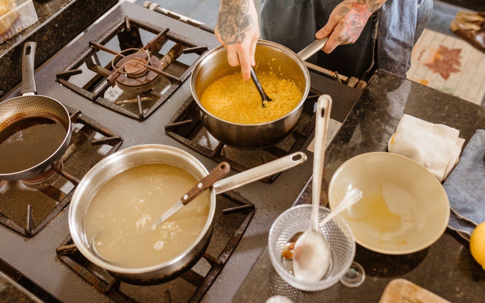 A cloth-tied pudding being lifted from a large stockpot with both hands, water dripping from the cloth, the pot on a gas hob