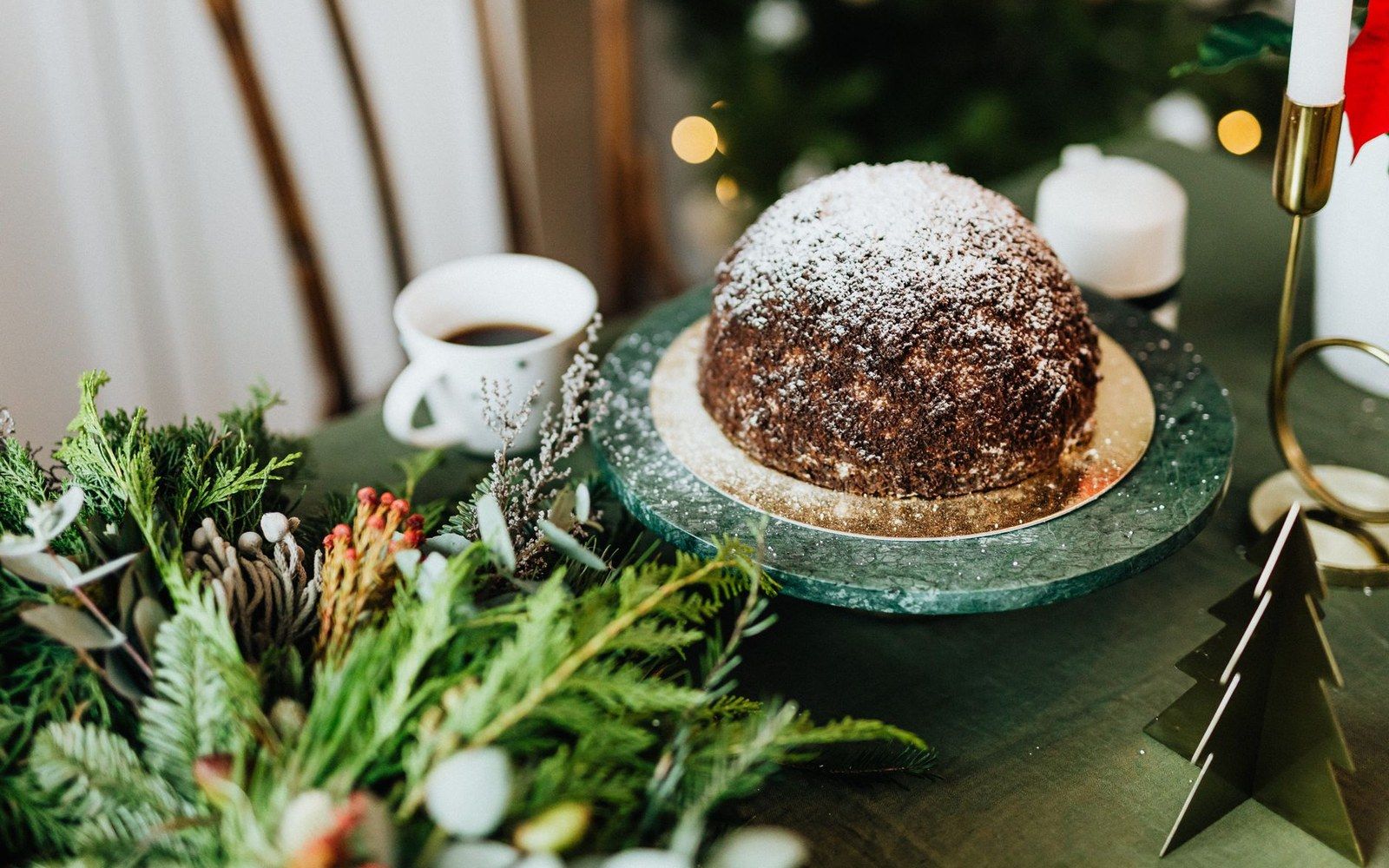 Domed christmas pudding dusted with powdered sugar on a marble stand beside coffee and greenery