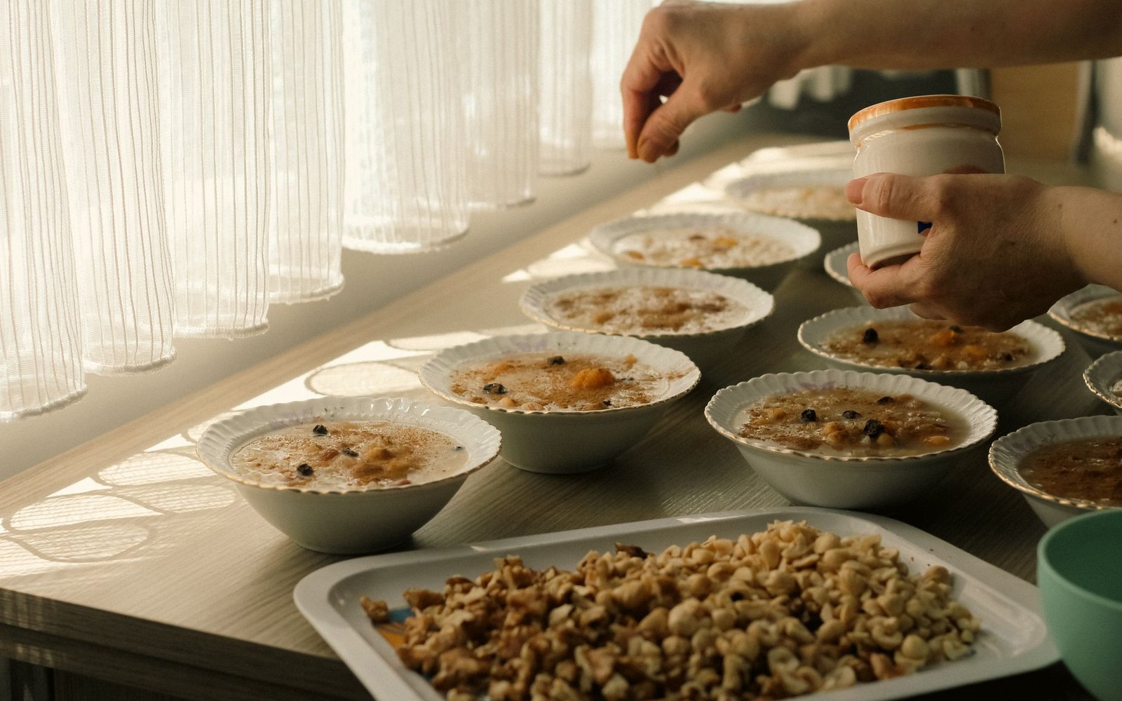 Hands sprinkle nuts from a jar onto rows of individual pudding bowls near a sunlit window