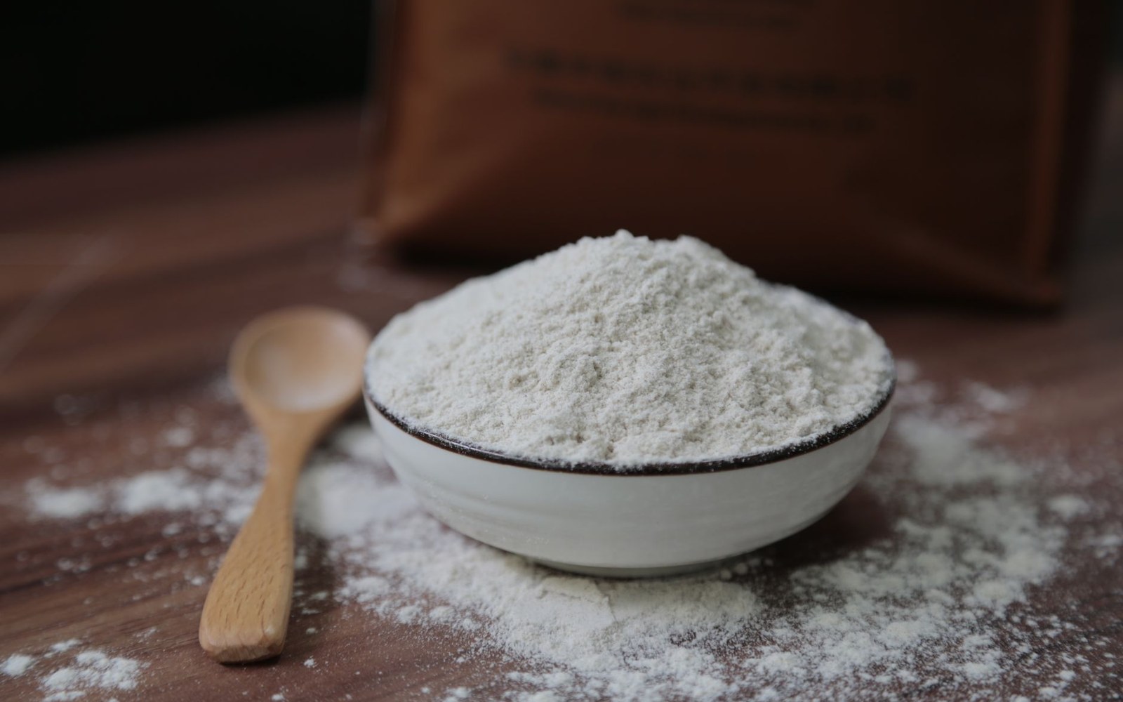 A bowl heaped with flour sits beside a wooden spoon on a scattered wooden table