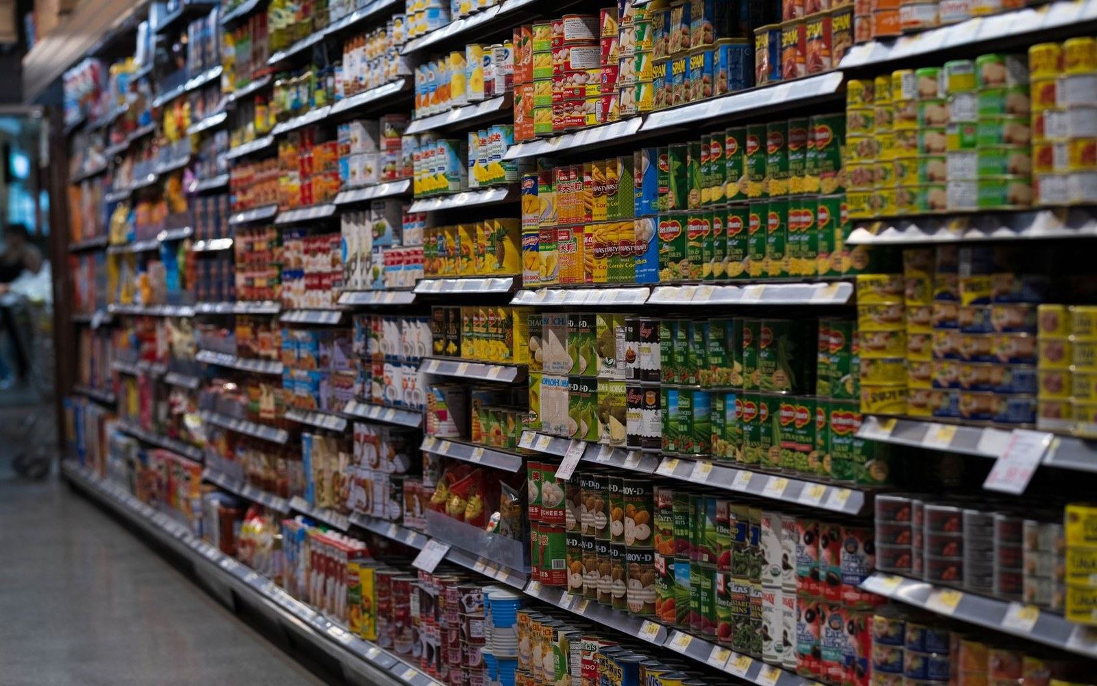 Rows of canned goods and packaged food line supermarket shelves with price tags below