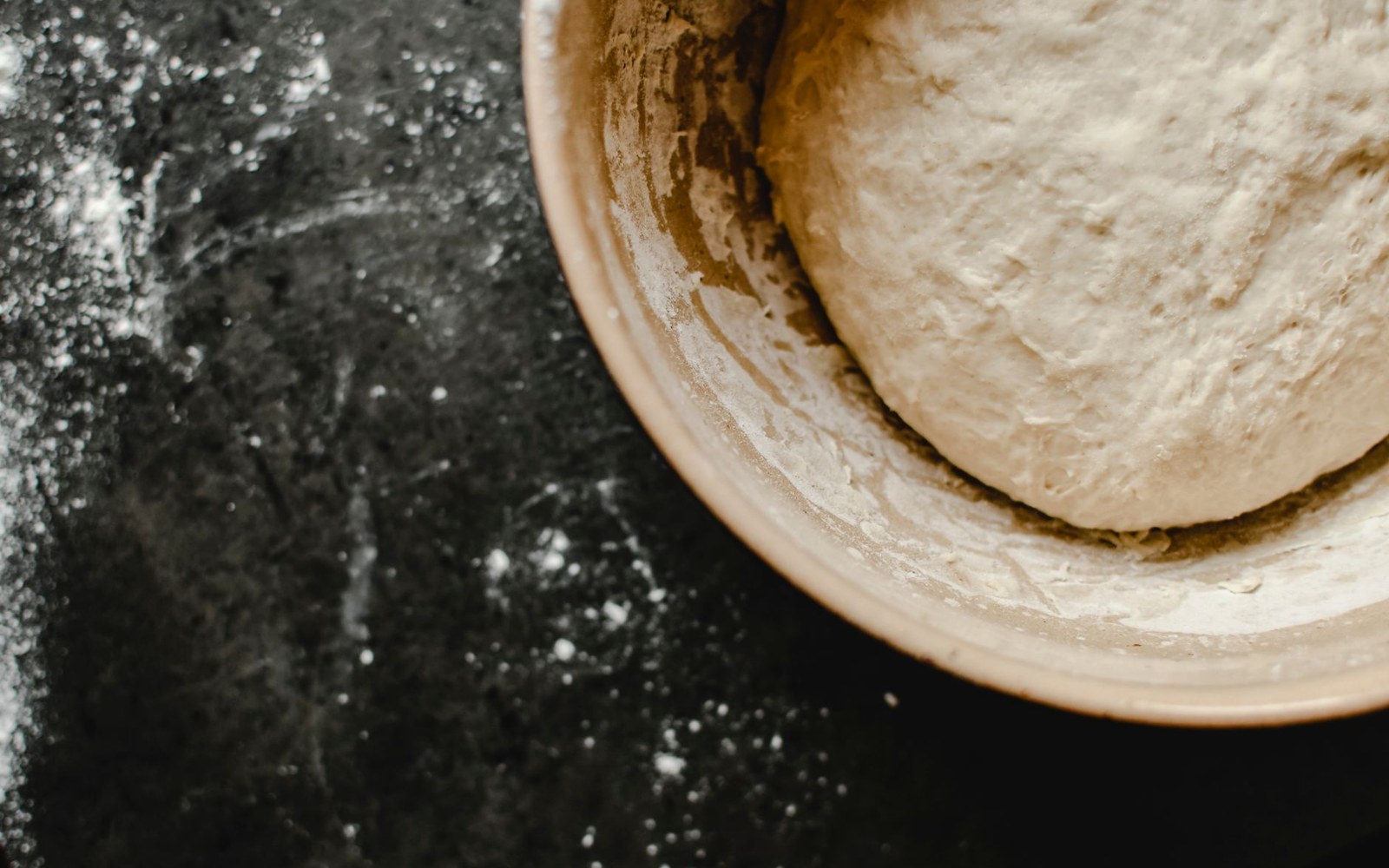 Dough rising in a ceramic bowl on a flour-dusted dark countertop