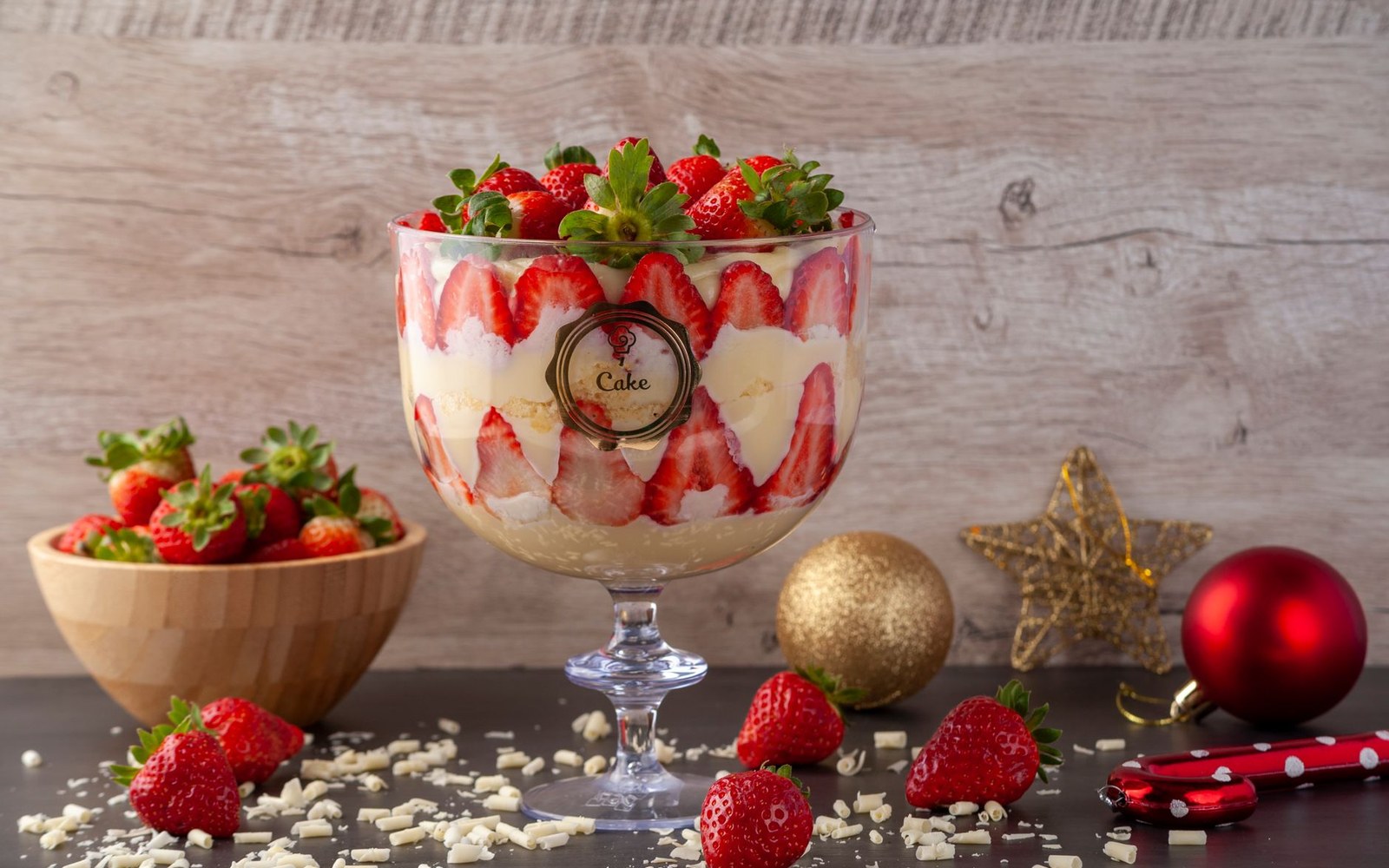 Layered strawberry trifle in a glass pedestal bowl surrounded by fresh strawberries and Christmas ornaments