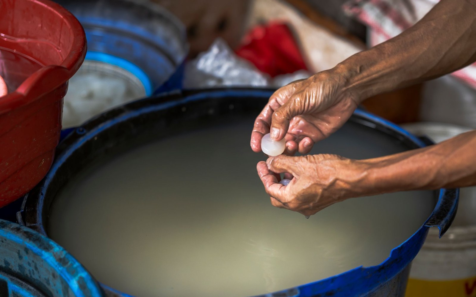 Adult hands pleating a sheet of greaseproof paper over the rim of a pudding basin, fingers folding the pleat, basin on a wooden surface