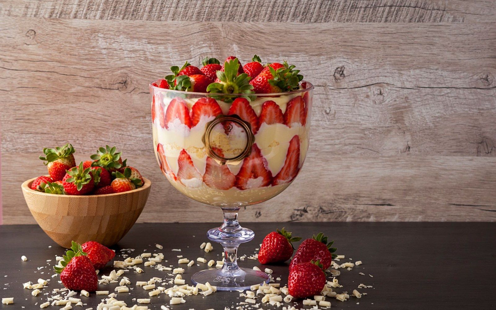 A glass trifle bowl on a kitchen counter showing distinct horizontal layers — sponge, custard, cream — shot from slightly above
